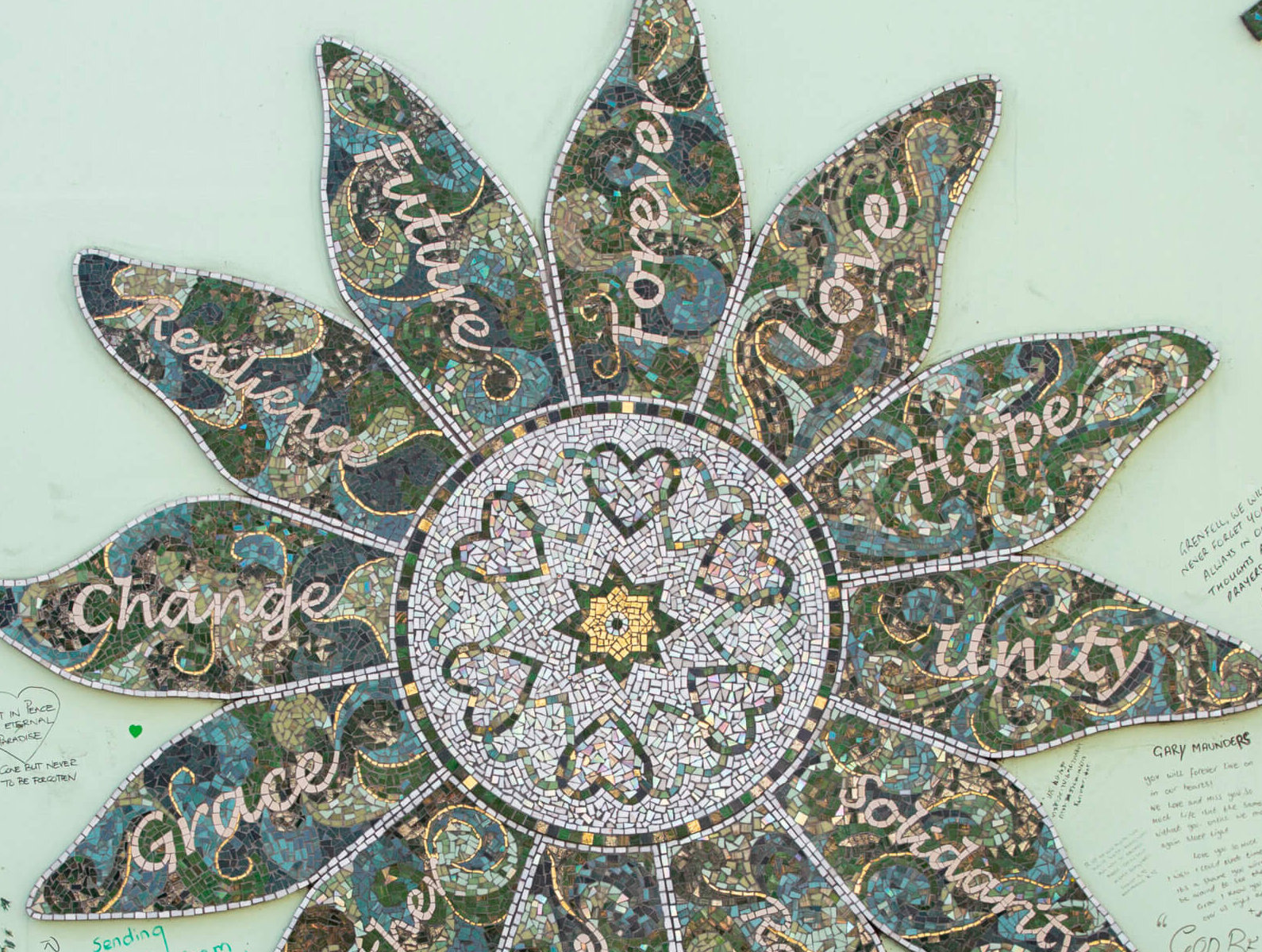 Grenfell flower image on wall, part of memeorial, with words like unity, future, love, hope, written on petals with colors in green and made in mosaics style.