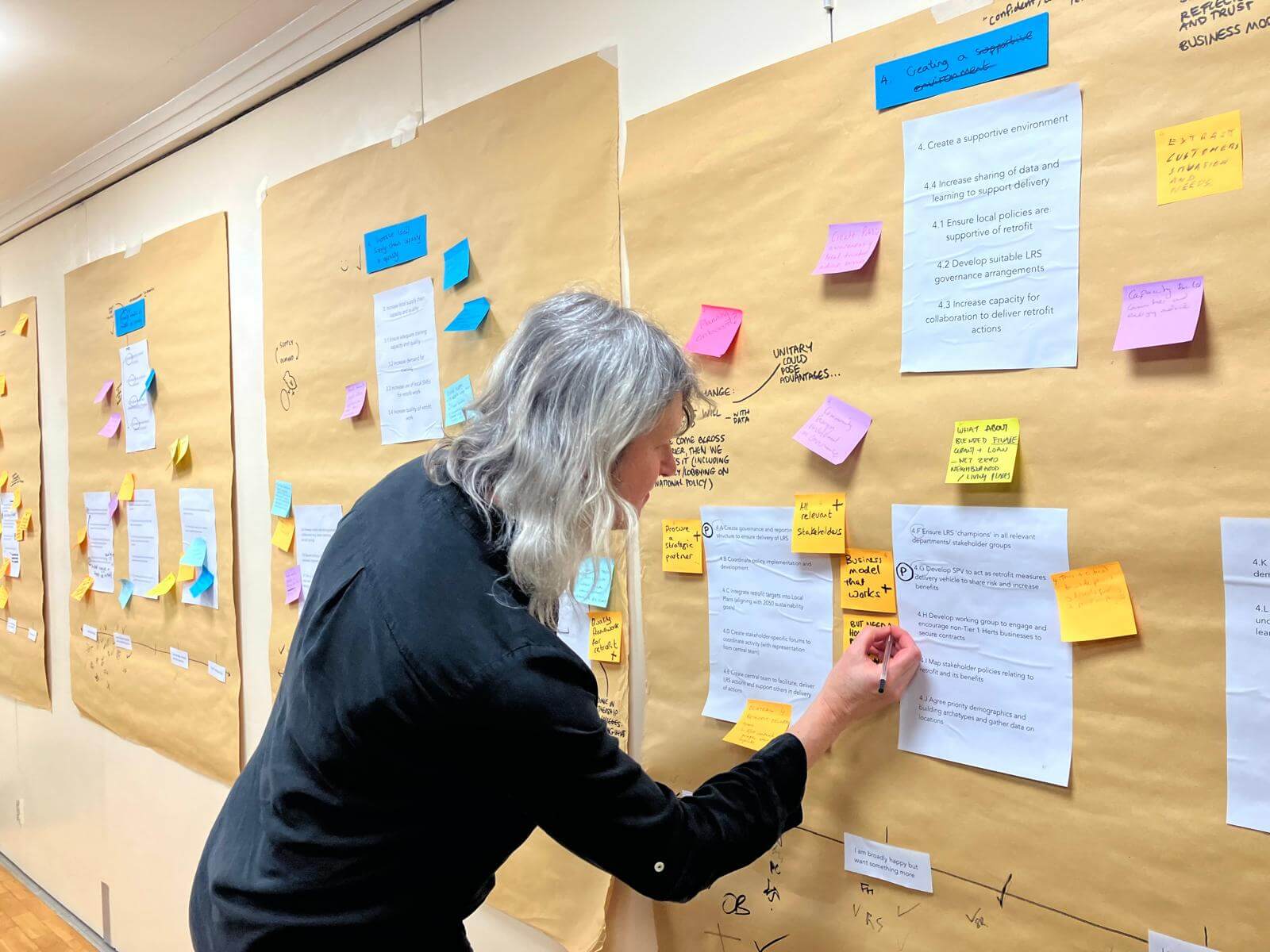 Image of Naomi standing at a board, with brown paper pinning up a post it note alongside lots of other multi coloured post its.